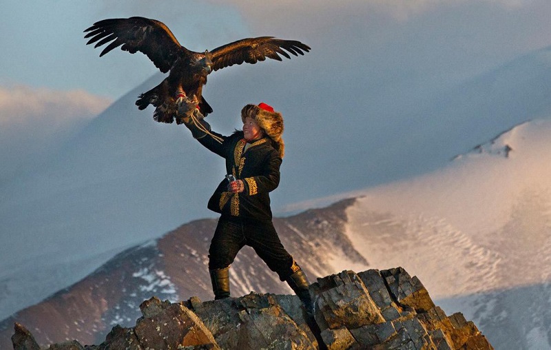 Aisholpan Nurgaiv đứng trên đồi núi với tay nâng cao một con đại bàng khổng lồ giữa không gian trong niềm kiêu hãnh của tuổi trẻ