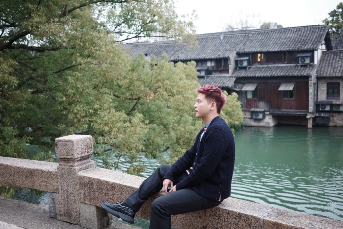 Anh chàng diện cardigan len tối màu phối cùng áo thun sáng bên trong và quần âu dáng suông. Sự tối giản màu sắc giúp tổng thể outfit trở nên tinh tế, phù hợp với không gian cổ kính, trầm lắng. Việc tiết chế màu sắc trong trang phục giúp Quân AP “hòa mình” vào bối cảnh mà vẫn giữ được nét hiện đại.